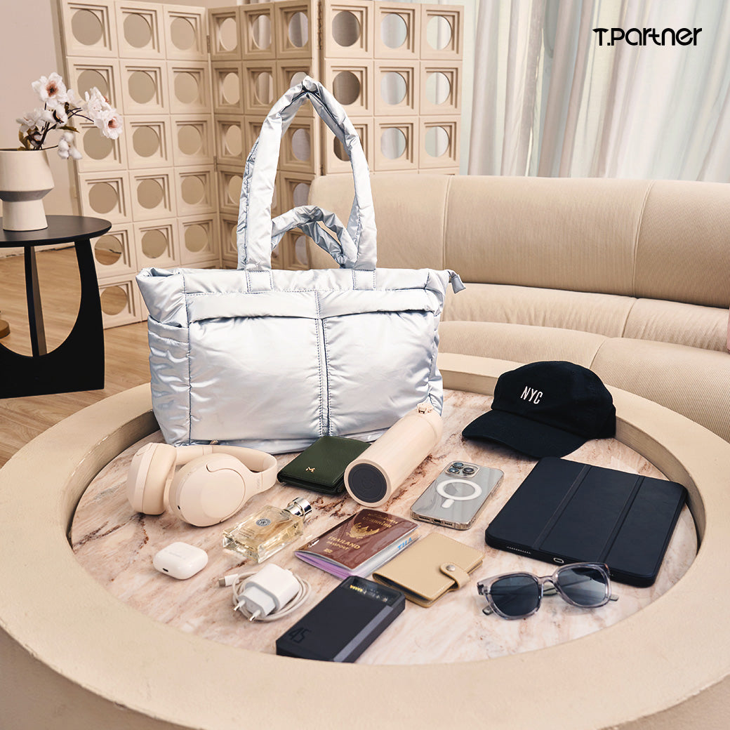 กระเป๋าสะพายขนาดใหญ่สีเงินเมทัลลิก Cozy Max bag พร้อมอุปกรณ์เดินทางอื่นๆ วางบนโต๊ะกลม